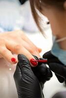 målning naglar av en kvinna. händer av manikyrist i svart handskar applicering röd nagel putsa på kvinna naglar i en skönhet salong. foto