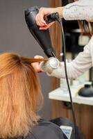 torkning hår i de hår studio. kvinna frisör frisör torkar hår med en hårtork och runda borsta röd hår av en kvinna i en skönhet salong. foto