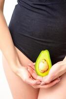 ung kvinna innehav ett halv av en färsk avokado frukt stänga till henne mage mot en vit bakgrund, friska näring och graviditet begrepp. foto