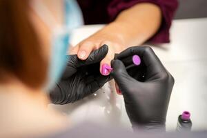 målning naglar av en kvinna. händer av manikyrist i svart handskar applicering rosa nagel putsa på kvinna naglar i en skönhet salong. foto