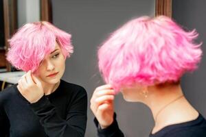 skön ung caucasian kvinna ser på henne kort rosa hår i en spegel. foto