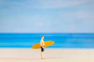 miniatyr- människor man i en baddräkt, och innehav en gul surfingbräda på de strand foto