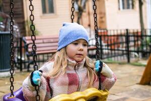 en ung flicka sitter på en gunga i en lekplats, henne uttryck är omtänksam och introspektiv. hon bär en mysigt, tillfällig utrusta, värma hatt och vantar. foto
