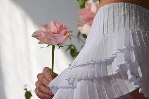 en brud flicka i en vit klänning innehar en delikat rosa reste sig blomma i henne hand. foto