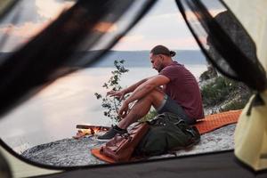 utsikt från resenärens tält med ryggsäck som sitter på toppen av berget och njuter av utsikten över en flod eller sjö. resa längs berg och kust, frihet och aktivt livsstilskoncept foto