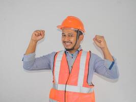 asiatisk man arbetstagare bär säkerhet hjälm lookis Lycklig fira hans seger förbi knäppning hans nävar mot grå bakgrund foto