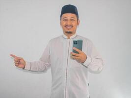 moslem asiatisk man leende Lycklig medan innehav mobil telefon och pekande till de rätt sida foto