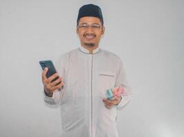 moslem asiatisk man leende Lycklig medan innehav mobil telefon och pengar foto
