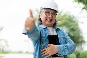 Lycklig äldre asiatisk professionell ingenjör bär en hjälm och checkar de plan på en läsplatta på de konstruktion webbplats efter de entreprenör och arkitekt Avsluta renovering de hus och byggnad foto