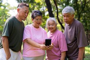 grupp av Lycklig senior pensionering använder sig av smartphone och skrattande utomhus på de parkera efter en träna och utgifterna tid tillsammans, begrepp handla om de äldre, anciennitet, och wellness åldring foto