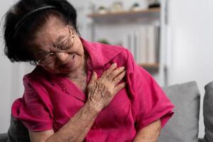 asiatisk senior äldre mormor lidande från hjärta smärta och innehav henne bröst medan Sammanträde på de soffa i henne levande rum. medicinsk nödsituation behandling och hälsa vård försäkring skydd begrepp foto