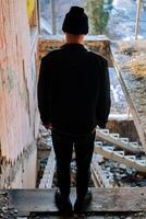 en stilig ung kille i en svart läder jacka står med hans tillbaka till de kamera mot de bakgrund av en förstörd trappa i ett oavslutat och övergiven byggnad. urban begrepp. vertikal skott foto
