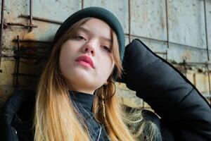porträtt av en skön Tonårs flicka i en svart vinter- jacka och hatt mot de bakgrund av ett oavslutat, förstörd och övergiven byggnad vägg. urban begrepp. närbild. foto