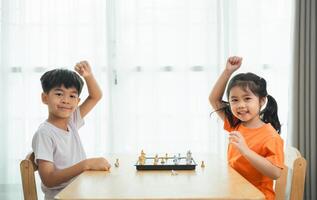 två barn spelar en spel av schack. ett av de barn är bär ett orange skjorta. de är både leende och verka till vara njuter de spel foto