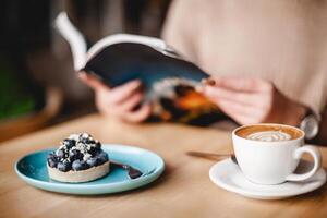 en kvinna njuter en lugn ögonblick, uppslukad i en bok medan njuter en blåbär syrlig och kaffe. de scen utstrålar avslappning och fritid, en härlig blandning av litteratur och kulinariska flathet foto