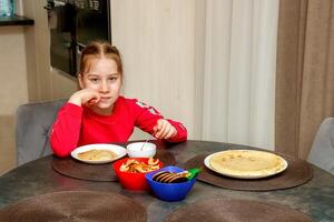 söt fundersam unge flicka på Hem på de tabell i främre av en tallrik av pannkakor foto
