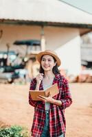 leende jordbrukare kvinna står innehav en anteckningsbok, klädd i en sugrör hatt och pläd skjorta, med en traktor i de bakgrund. foto