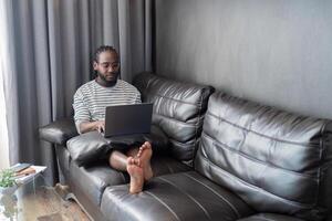 ung afrikansk amerikan man innehav bärbar dator avkopplande Sammanträde på soffa arbetssätt avlägsen på Hem foto