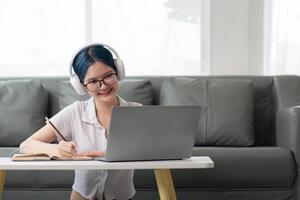 Lycklig ung kvinna Tonårs bär hörlurar skrivning notera. studerande uppkopplad inlärning klass studie uppkopplad ring upp zoom lärare med bärbar dator och bok foto