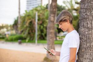 stänga upp stilig tonåring pojke i en vit t-shirt använder sig av mobil telefon utomhus i en parkera lunch tid, rum för text.sida se. foto
