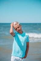 stilig tonåring pojke av europeisk utseende med blond hår i vit shorts, och en blå t-shirt står i de hav i de vatten, och utseende till de kamera. sommar semester koncept.snygg tonåring pojke porträtt koncept.vertikal Foto. foto