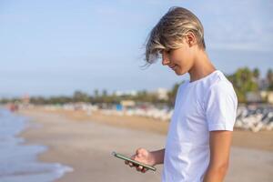 stänga upp stilig tonåring pojke i vit t-shirt använder sig av smartphone på de strand på sommar högtider, resa eller resa i Spanien .resa,semester,semester, frihet koncept.sida se. foto