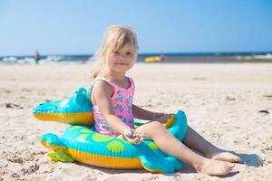 Lycklig flicka av europeisk utseende ålder av 5 Sammanträde på ett uppblåsbar krokodil leksak på de strand sommar solig dag, ser till de kamera.familj sommar yrke begrepp. kopia Plats. foto