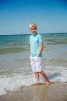 stilig tonåring pojke av europeisk utseende med blond hår i vit shorts, och en blå t-shirt står i de hav i de vatten, och utseende till de kamera. sommar yrke koncept.sommar resa försäljning koncept.vertikal Foto. foto