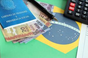 brasiliansk arbete kort och social säkerhet blå bok och reais pengar räkningar med kalkylator och penna på flagga av federativ republik av Brasilien foto