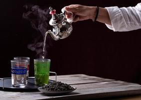 person som serverar te i en silvertekanna över exotiska färgade glasvaser foto