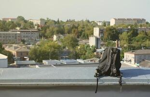 svart ryggsäck lögner på metall gräns av bosatt flervåningshus byggnad taket i solig väder foto