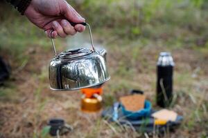 metall vattenkokare tillverkad av rostfri stål, camping redskap, vandring Utrustning, varm te, koka upp vatten i natur, hand innehar vattenkokare med kokande vatten. frukost av en turist i de skog. foto