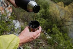 en hand innehar en råna av te, vatten häller från ovan från en termos, en turist på en vandra häller en varm dryck in i en glas, en termo flaska, en svart termos, maträtter i en camping i natur. foto