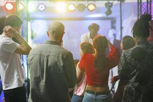 ung människor stående på dansgolv medan delta elektronisk musik konsert i nattklubb. olika män och kvinnor folkmassan dans medan dj utför på skede med färgrik spotlights foto