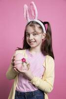 Lycklig positiv flicka med kanin öron innehar en rosa påsk ägg i studio, känsla stolt handla om henne festlig dekorationer handgjorda. små barn varelse självsäker på kamera, vår Semester händelse. foto