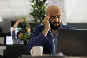 trött arab affärsman med huvudvärk arbetssätt i företag kontor. överarbetad verkställande chef lidande från migrän och gnuggning tempel medan analyserar Start upp företag Rapportera på bärbar dator foto