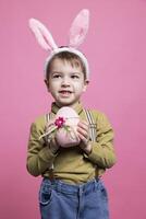 glad ung barn som visar en rosa ägg dekorerad för påsk, känsla Lycklig och upphetsad handla om vår Semester händelse. liten söt unge innehav handgjort festlig prydnad, bär kanin öron i studio. foto