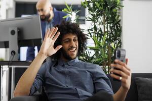 Lycklig arab affärsman vinka Hej medan talande på smartphone uppkopplad möte i Start upp företag kontor. företag verkställande chef hälsning avlägsen medarbetare på mobil telefon ring upp foto