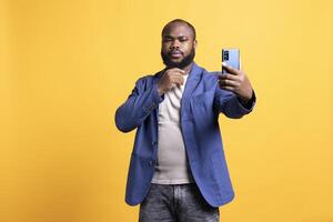 narcissistisk man använder sig av mobiltelefon till ta selfies, strök hans haka. fåfäng social media användare tar foton använder sig av telefon selfie kamera, isolerat över gul studio bakgrund