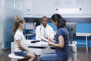 fyllning ut pappersarbete för en medicinsk examen på de sjukhus kontor med henne unge är en caucasian mor. medan de kvinna fyllningar ut papper på de klinik ett afrikansk amerikan läkare väntar. foto