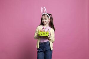 Lycklig självsäker flicka med kanin öron presenter en korg fylld med målad handgjort påsk dekorationer över rosa bakgrund. ung glad unge som visar festlig färgrik ornament i studio. foto