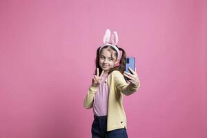 glad Lycklig barn tar bilder och som visar fred tecken på telefon, känsla glad och självsäker handla om roligt foton för påsk firande. liten skol med pigtails tar fotografier.