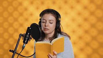 berättare bär headsetet läsning högt från bok in i mic mot gul bakgrund. optimistisk professionell röst skådespelare inspelning ljudbok, skapande engagerande media innehåll för lyssnare, kamera en foto