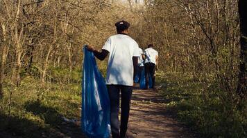 afrikansk amerikan flicka samlar skräp i en skräp väska använder sig av tång, återvinning plast avfall och plockning upp sopor i en skog. ung kvinna aktivist clearing de trän från skräp. kamera b. foto