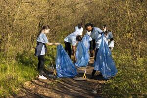 grupp av olika volontärer samlar skräp och plast avfall, använder sig av påsar till återvinna och samla Allt skräp från de skog livsmiljö. aktivister samarbetande till klar de trän område från skräp. foto