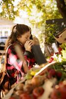 selektiv fokus av ung vuxen lukta nyligen UPPTAGITS äpple på eco vänlig marknad. kvinna shopper välja och köp av lokalt vuxen organisk bio frukt och grönsaker på utomhus- mat marknadsföra. foto