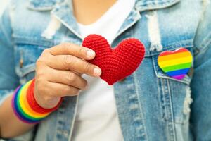 asiatisk dam som bär regnbågsflaggarmband och håller rött hjärta, symbol för HBT stolthet månad firar årliga i juni sociala av homosexuella, lesbiska, bisexuella, transpersoner, mänskliga rättigheter. foto
