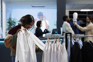 afrikansk amerikan kvinna påfrestande på skjorta, kontroll storlek och passa medan handla i Kläder Lagra. kund granskning kläder medan välja formell utrusta i köpcenter mode boutique foto