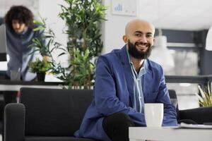 leende arab företag entreprenör ser på kamera medan Sammanträde på soffa i kontor. Start upp företag framgångsrik verkställande chef bär kostym porträtt i coworking Plats foto