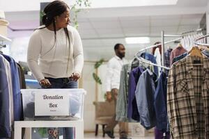 afrikansk amerikan kvinna välja kläder till sätta i donation låda i handla Centrum. mode boutique klient donera ny och Begagnade apprel i plast behållare för humanitär hjälpa foto